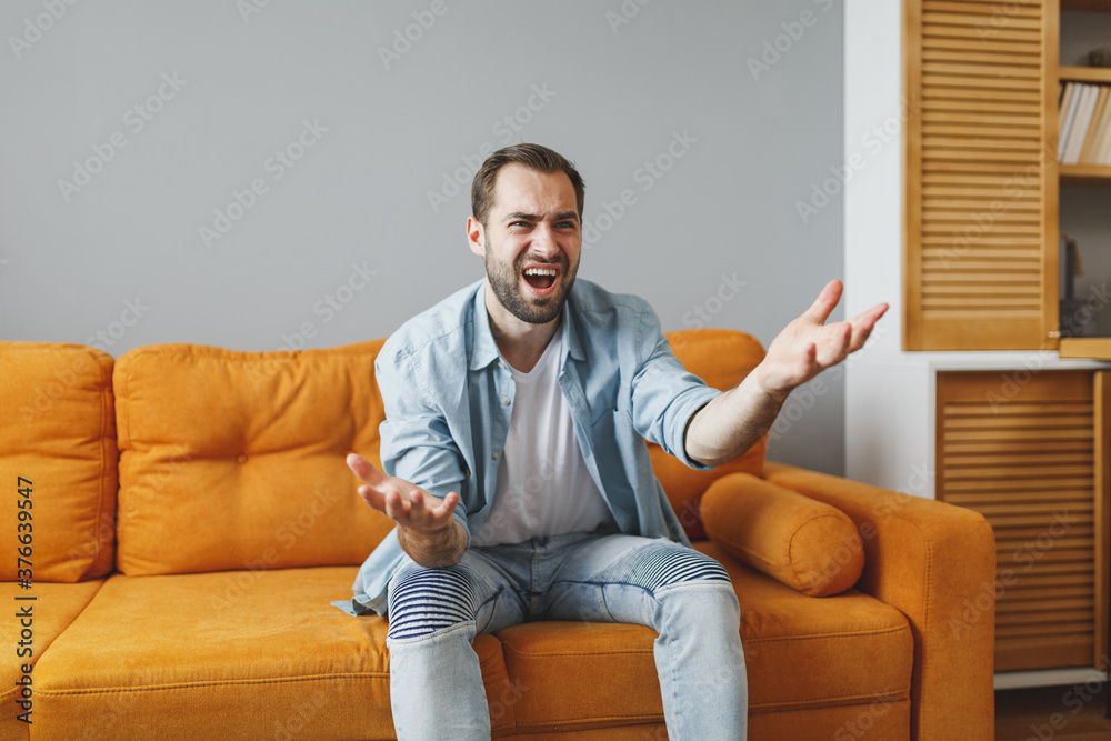Displeased irritated young bearded man 20s wearing casual white t-shirt blue shirt looking aside swearing spreading hands sitting on couch resting relaxing spending time in living room at home.