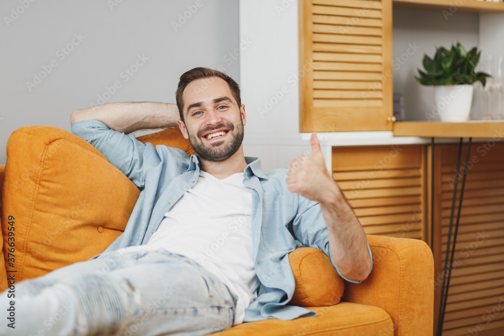 Smiling cheerful handsome attractive young bearded man wearing casual white t-shirt blue shirt looking camera showing thumb up lying on couch resting relaxing spending time in living room at home.