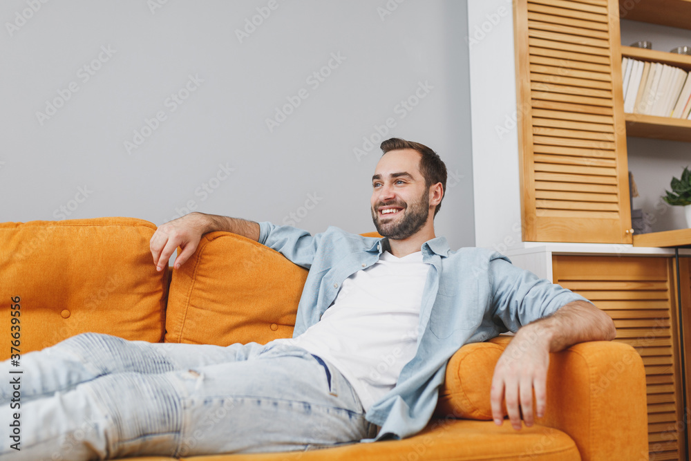 Smiling handsome attractive pleasant young bearded man 20s wearing casual white t-shirt blue shirt looking aside sitting on couch resting relaxing spending time in living room at home. Smiling handsome attractive pleasant young bearded man 20s wearing casual white t-shirt blue shirt looking aside sitting on couch resting relaxing spending time in living room at home.