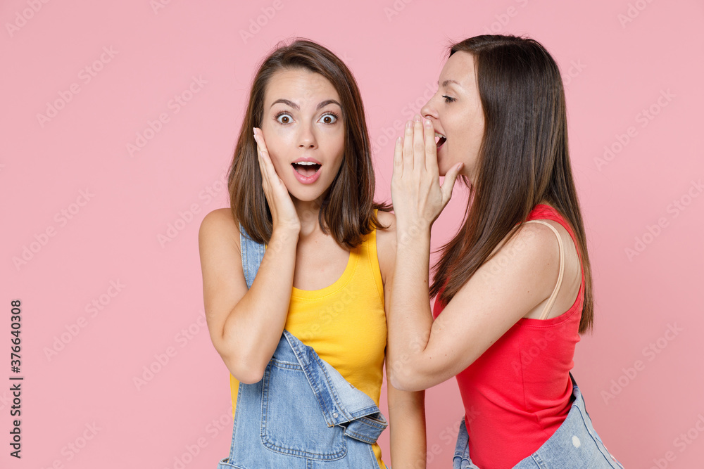 Φωτογραφία Two shocked amazed young women friends 20s in casual denim clothes posing whispering secret behind hand, sharing news put palm on cheek isolated on pastel pink colour background, studio portrait