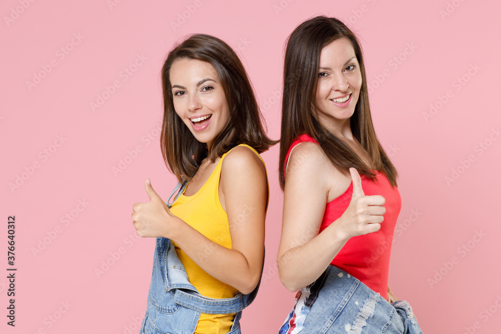 Side view of two cheerful smiling young women friends 20s in casual denim clothes standing back to back showing thumbs up looking camera isolated on pastel pink colour background, studio portrait. Side view of two cheerful smiling young women friends 20s in casual denim clothes standing back to back showing thumbs up looking camera isolated on pastel pink colour background, studio portrait.