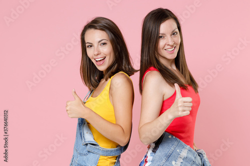Side view of two cheerful smiling young women friends 20s in casual denim clothes standing back to back showing thumbs up looking camera isolated on pastel pink colour background, studio portrait.