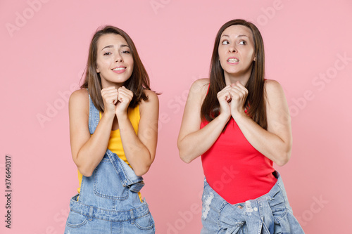 Two excited worried pleading young women friends 20s wearing casual denim clothes posing clenching fists wait for special moment making wish isolated on pastel pink colour background, studio portrait.