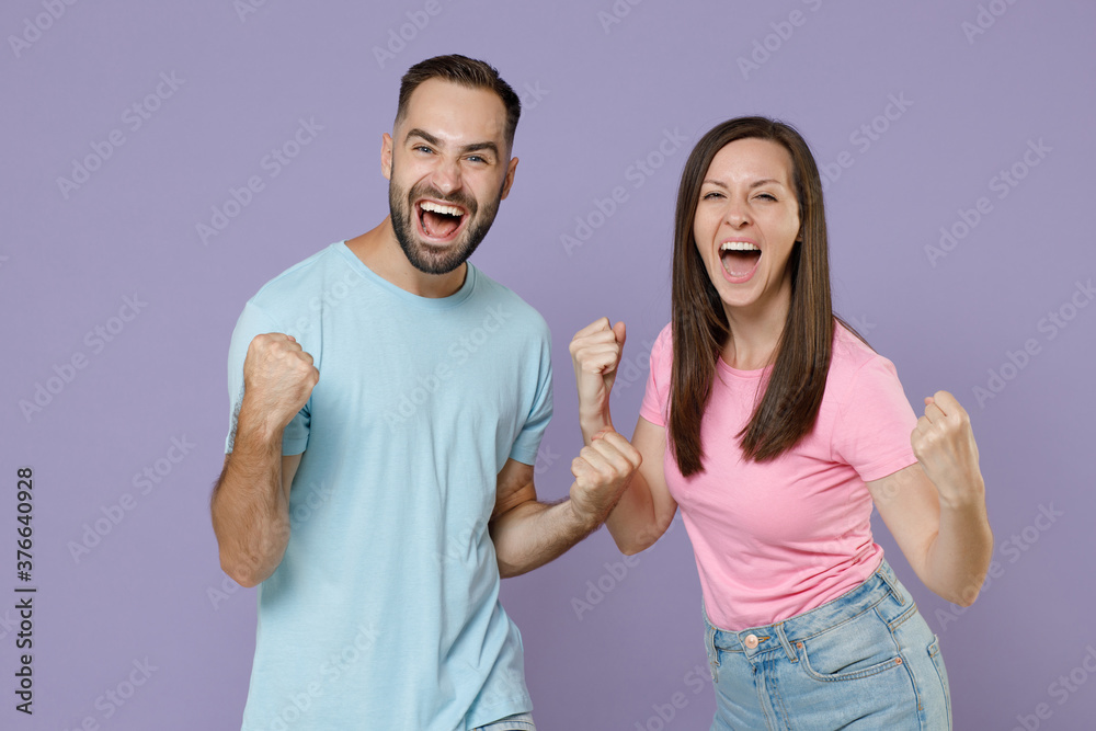 Excited happy joyful young couple two friends man woman 20s in blue pink empty blank design t-shirts clenching fists doing winner gesture isolated on pastel violet color background studio portrait. Excited happy joyful young couple two friends man woman 20s in blue pink empty blank design t-shirts clenching fists doing winner gesture isolated on pastel violet color background studio portrait.