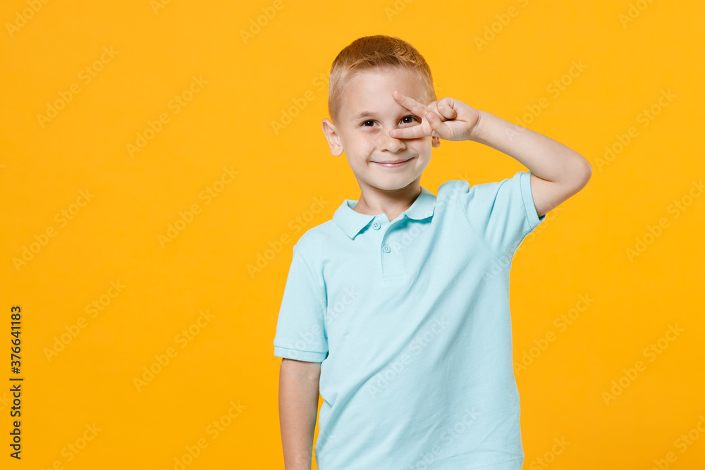 Little fun male fair-haired brown-eyed kid boy 5-6 years old wearing stylish blue turquoise t-shirt polo showing victory sign near eye isolated on yellow color wall background, child studio portrait. Little fun male fair-haired brown-eyed kid boy 5-6 years old wearing stylish blue turquoise t-shirt polo showing victory sign near eye isolated on yellow color wall background, child studio portrait.