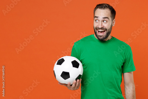 Excited young man football fan in green t-shirt cheer up support favorite team with soccer ball keeping mouth open looking aside isolated on orange background. People sport leisure lifestyle concept.