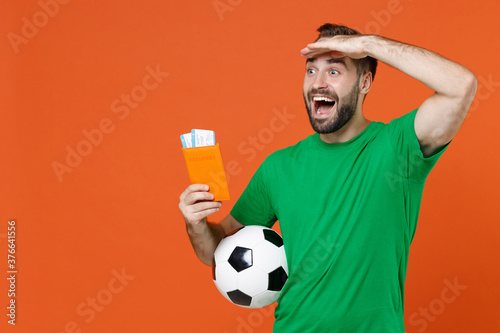 Excited man football fan in green t-shirt cheer up support favorite team with soccer ball hold passport ticket looking far away distance isolated on orange background. People sport leisure concept.