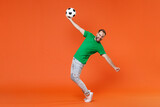 Full length portrait funny man football fan in green t-shirt cheer up support favorite team with soccer ball spreading hands stand on toes isolated on orange background. People sport leisure concept.