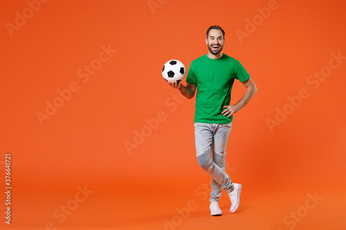 Fototapeta Naklejka Na Ścianę i Meble -  Full length portrait funny young man football fan in basic green t-shirt cheer up support favorite team with soccer ball isolated on orange background studio. People sport leisure lifestyle concept.