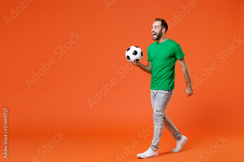 Fototapeta Naklejka Na Ścianę i Meble -  Full length portrait cheerful young man football fan in basic green t-shirt cheer up support favorite team with soccer ball looking aside isolated on orange background. People sport leisure concept.