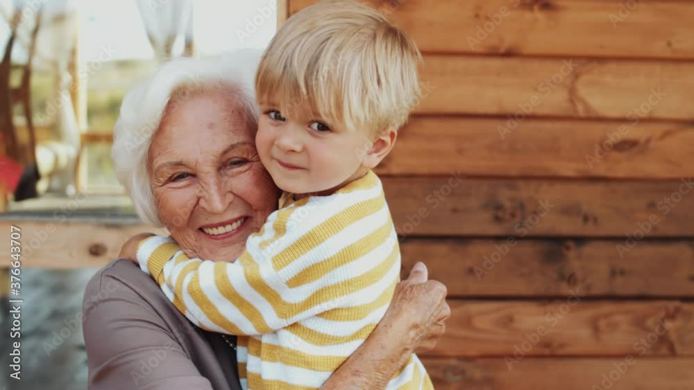 Adorable little boy and his senior grandmother hugging each other, looking at camera and smiling while posing for camera on wooden terrace on summer day Adorable little boy and his senior grandmother hugging each other, looking at camera and smiling while posing for camera on wooden terrace on summer day