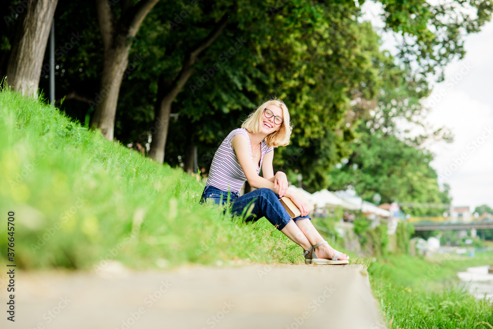 Literature for summer vacation. Girl relaxing at riverside after working day. Rest relax and hobby. Summer vacation. Girl sit on green grass near river and read book. Woman dreaming about vacation Literature for summer vacation. Girl relaxing at riverside after working day. Rest relax and hobby. Summer vacation. Girl sit on green grass near river and read book. Woman dreaming about vacation