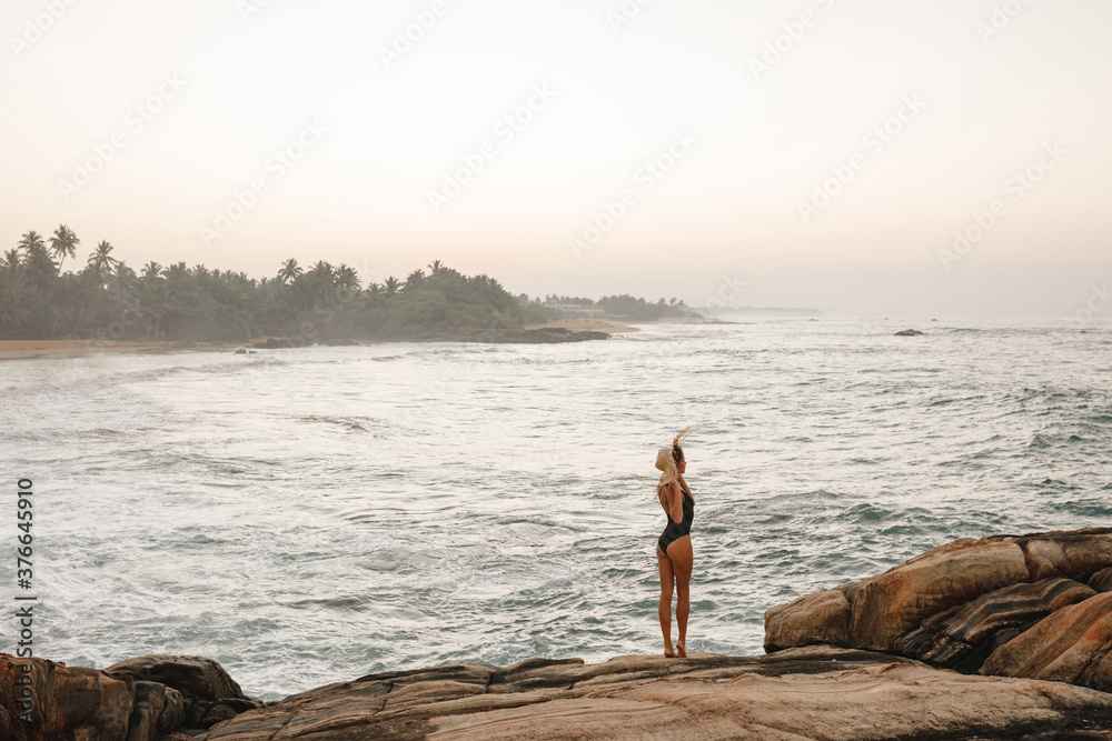 Back view young woman in black swimsuit and straw hat stand on rocky seashore and enjoy beautiful sea view