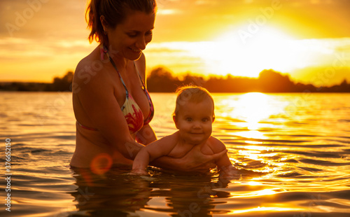 Side view of happy caucasian mother swimming with little cute smiling baby in sea. Beautiful family enjoying summertime outdoors during fantastic bright orange sunset. Family concept. © YouraPechkin