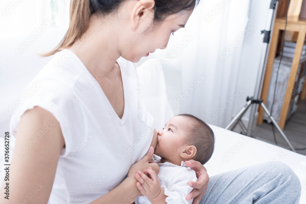 Mother breastfeeding her newborn baby beside window. Milk from mom’s breast is a natural medicine for children. Woman feeding baby. A Young mother holding her baby child.