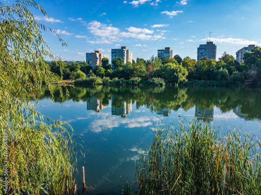 City landscape. Residential buildings are reflected in the water of a lake with green vegetation along the shore on a sunny day under a blue sky with branches in the foreground.