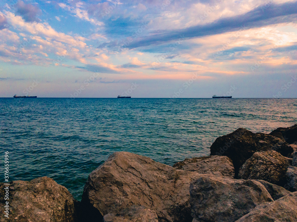 The surface of the sea against a blue cloudy sky with three ships on the horizon and large stones in the foreground.
