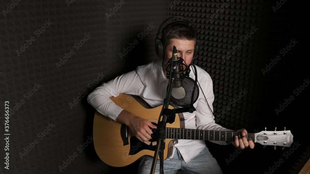 Musician recording new song in a modern sound studio. Young man dressed in a white shirt is singing and playing on a guitar in studio. Black wall in background. Musician recording new song in a modern sound studio. Young man dressed in a white shirt is singing and playing on a guitar in studio. Black wall in background.