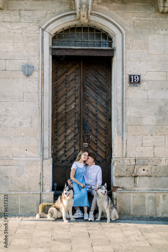 Happy couple with two husky dogs having fun outdoors in the city, sitting near ancient wooden door in front of old stone building. Full length photo
