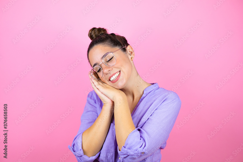 Young beautiful woman wearing glasses over isolated pink background sleeping tired dreaming and posing with hands together while smiling with closed eyes.