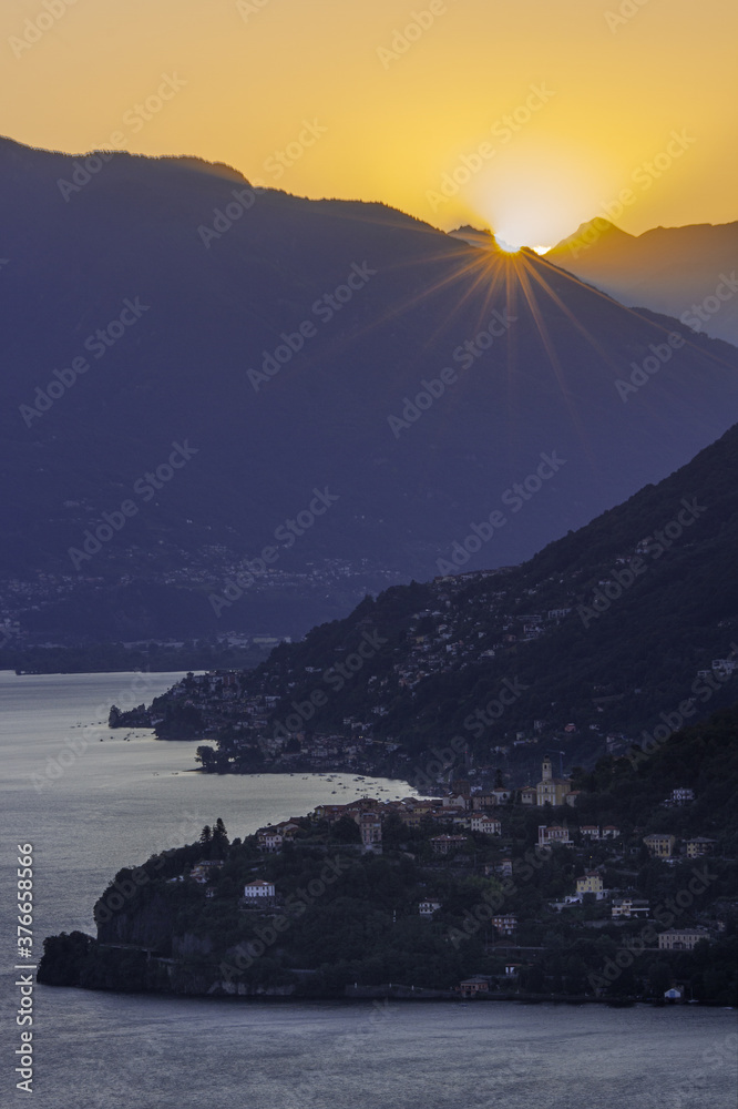 Il sole sorge spuntando dalle vette che circondano il Lago Maggiore illuminando il paese di Pino sulla sponda del Lago Maggiore
