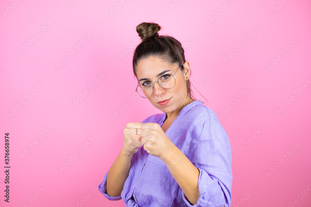 Young beautiful woman wearing glasses over isolated pink background Punching fist to fight, aggressive and angry attack, threat and violence Young beautiful woman wearing glasses over isolated pink background Punching fist to fight, aggressive and angry attack, threat and violence