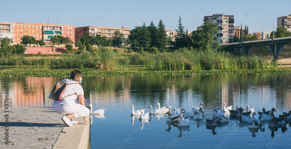 Obraz premium Rear View Of A Beautiful Young Girl On A Jetty Looking At The Ducks In The River. Woman with a protective mask due to Covid-19.