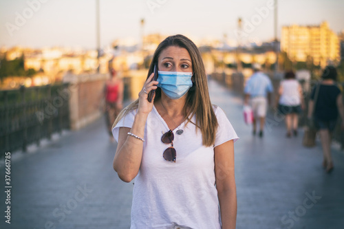 Woman Wearing Face Mask Using Phone.   Attractive woman walking through crowed street wearing protective face mask due to covid-19. She is using her phone. © Nanci