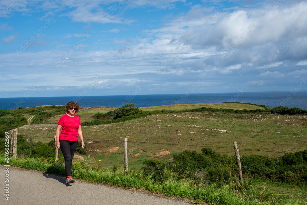 Mature woman dressed in sportswear walking along a country road with the sea in the background, horizontal