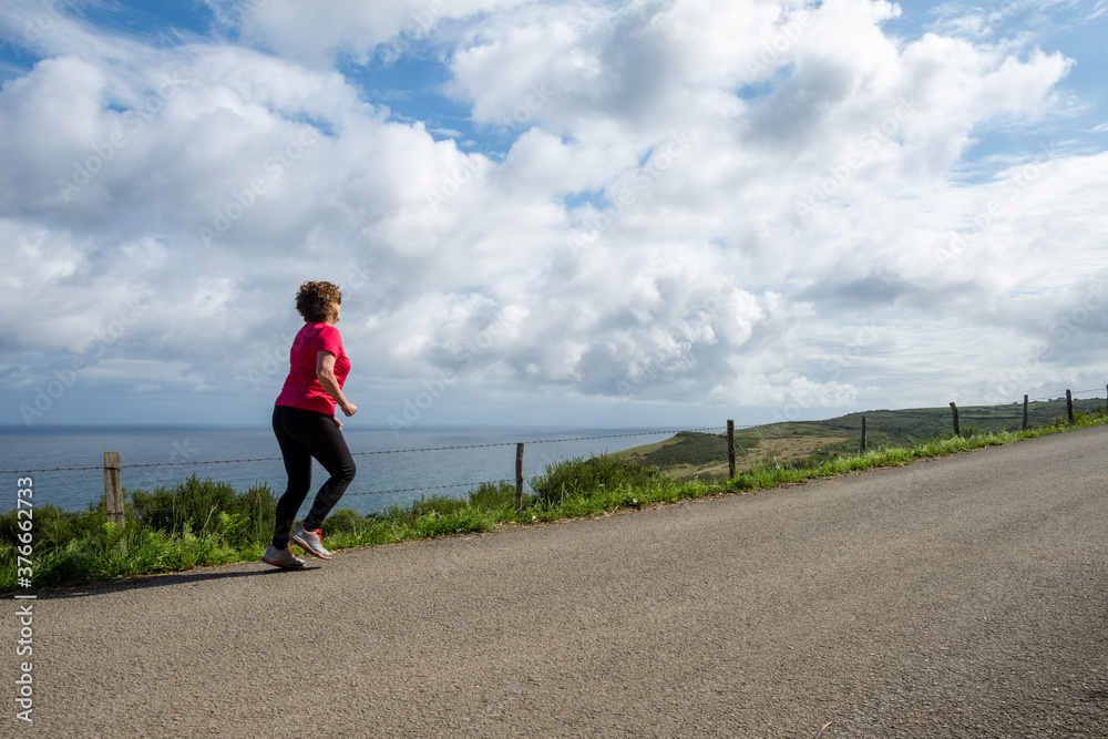 General shot of mature woman wearing sportswear, running on a rural road, a bright day against a background of sky with clouds, horizontally, with copy space