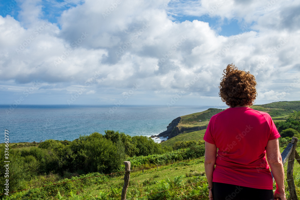 Mature woman with sportswear, from behind, looking at a landscape of field and sea, a bright day, in Cantabria, Spain, horizontal, with copy space