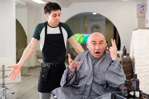 Portrait of angry man with shaved head shocked by haircut and regretting hairdresser in modern barbershop