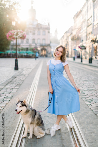 Full length summer outdoor portrait of young attractive girl in blue dress, posing with her husky dog, standing on tram track on the background of old buildings in European city