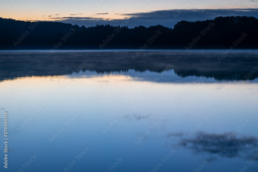 Fototapeta premium Cool and cloudy morning at peaceful and romantic lake Ukleisee with forest in background and smooth water in foreground, Eutin, Schleswig-Holstein, Northern Germany