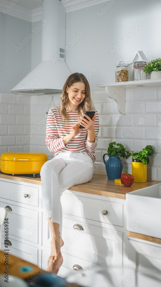 Beautiful Young Female in Striped Jumper and White Pants is Sitting on a Kitchen Furniture and Using Her Smartphone a Modern Sunny Kitchen. She is Happy and Excited while Browsing Social Media. Beautiful Young Female in Striped Jumper and White Pants is Sitting on a Kitchen Furniture and Using Her Smartphone a Modern Sunny Kitchen. She is Happy and Excited while Browsing Social Media.