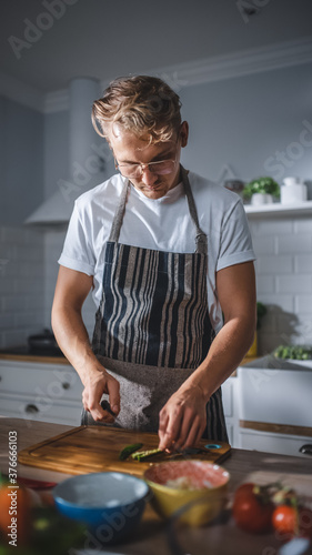 Handsome Man in White Shirt and Apron is Making a Healthy Organic Salad Meal in a Modern Sunny Kitchen. Hipster Man in Glasses Cooking. Natural Clean Diet and Healthy Way of Life Concept.