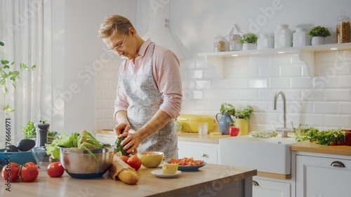 Handsome Man in Pink Shirt and Apron is Making a Healthy Organic Salad Meal in a Modern Sunny Kitchen. Hipster Man in Glasses. Natural Clean Diet and Healthy Way of Life Concept.