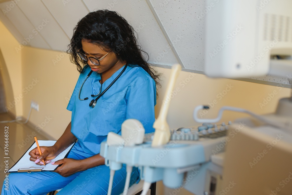 Obraz premium Young cheerful African woman operator of an ultrasound scanning machine analyzing diagnostics results of patient. Young smiling African doctor working on a modern ultrasound equipment.