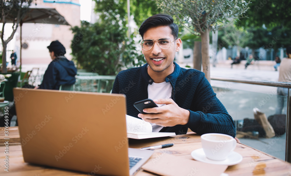 Positive young male with smartphone and laptop enjoying coffee in street cafe Positive young male with smartphone and laptop enjoying coffee in street cafe