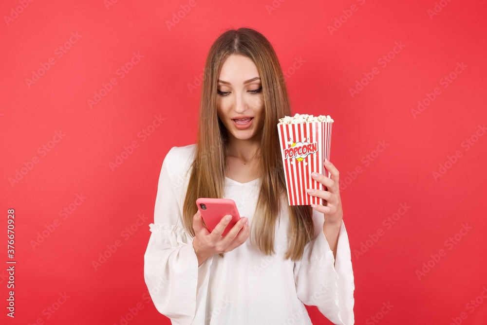Eastern European woman holding bucket with popcorn standing over isolated red background holding in hands cell reading sms using new app 5g