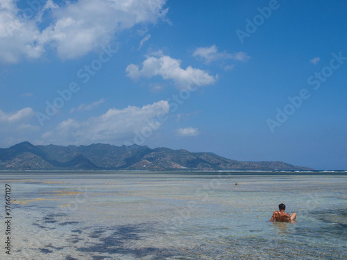 Paradaise beach in blue tones with turquoise water, mountains at the background and a man on his back swimming in the sea photo