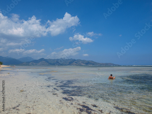 Paradaise beach in blue tones with turquoise water, mountains at the background and a man on his back swimming in the sea photo