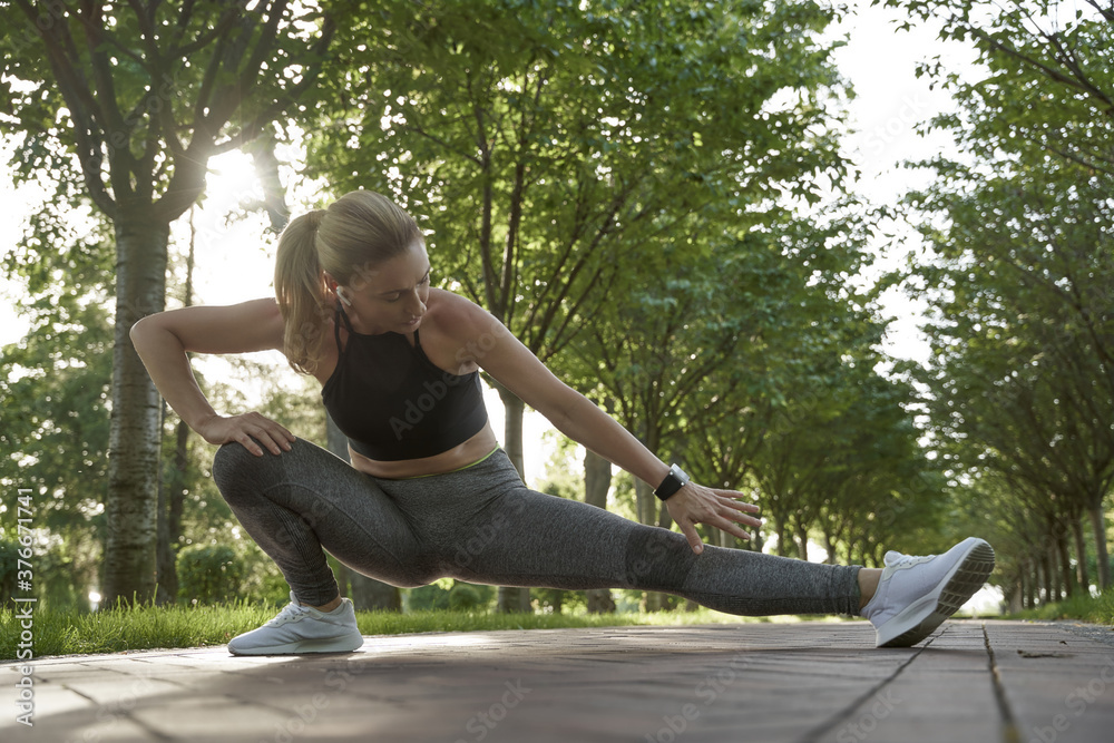 Fototapeta premium Warming up outdoors. Full length of beautiful fitness woman in sportswear stretching her legs while exercising in city park on a sunny summer morning