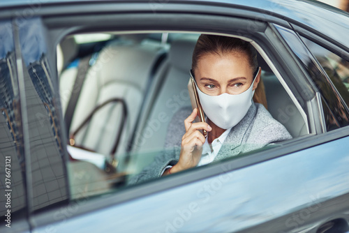 Covid 19 or coronavirus outbreak, caucasian businesswoman wearing medical protective mask talking on smartphone while sitting on back seat in the car, looking out of a window