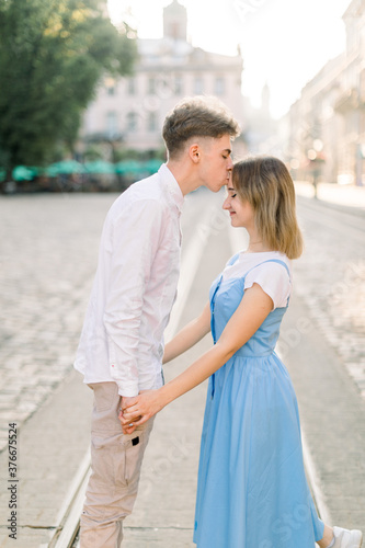 Handsome young man kissing his charming pretty girlfriend on forehead in the street in the sunny morning. Romantic couple in the street in ancient European city