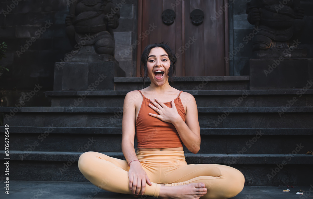 Portrait of expressive caucasian female in active wear enjoying free time and practice surprised with something, funny teen cheerful woman sitting in lotus pose smiling and looking at camera