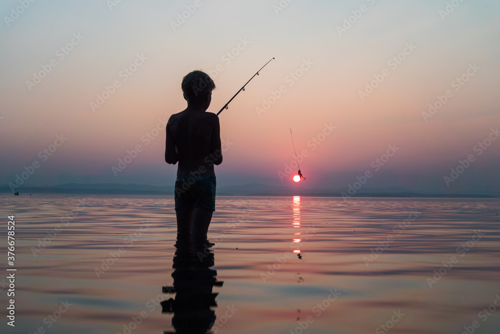 boy fishing with a fishing rod standing in the water in the evening at sunset