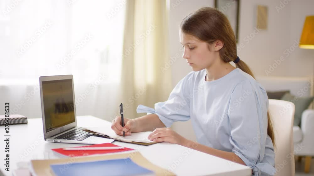Medium shot of cheerful schoolgirl sitting at her desk and writing in notebook while talking to teacher on video call, then waving goodbye, closing laptop and stretching Medium shot of cheerful schoolgirl sitting at her desk and writing in notebook while talking to teacher on video call, then waving goodbye, closing laptop and stretching