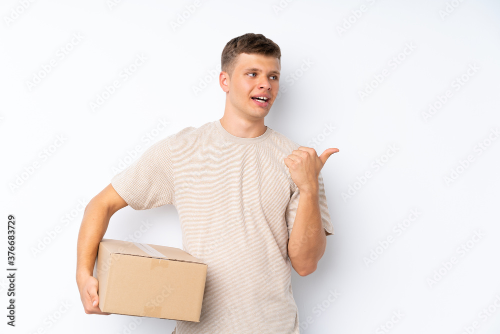 Young handsome man over isolated white background holding a box to move it to another site and pointing side Young handsome man over isolated white background holding a box to move it to another site and pointing side