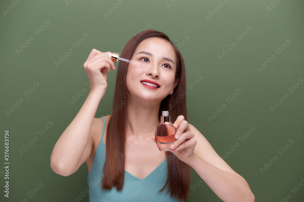 Obraz premium Close up asian woman 20s with perfect skin, nude make up apply oil from bottle on face isolated on green wall background